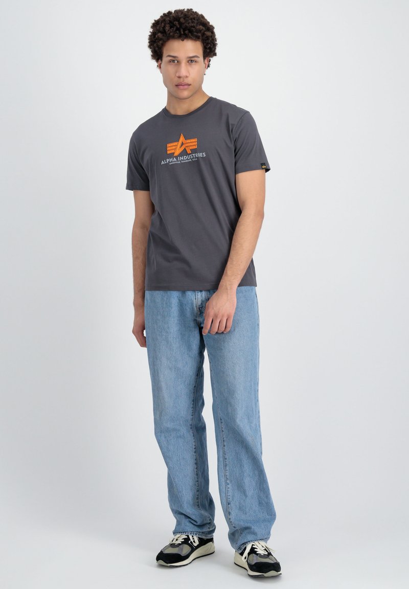 Graues Baumwoll-T-Shirt mit kurzen Ärmeln, versehen mit einem orangen Logo, kombiniert mit hellblauen Jeans und schwarzen Sneakers. Einfaches Design und Passform.