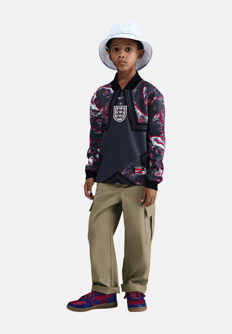 Junger Junge trägt ein England Fußballtrikot, beige Cargohosen, rot-blaue Sneaker und einen weißen Fischerhut und steht vor einem schlichten Hintergrund.