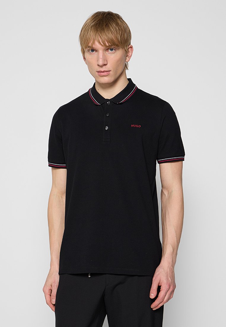 Blonder Mann trägt ein schwarzes Hugo-Poloshirt mit roten und weißen Akzenten an Kragen und Ärmeln und steht vor einem schlichten, hellen Hintergrund.