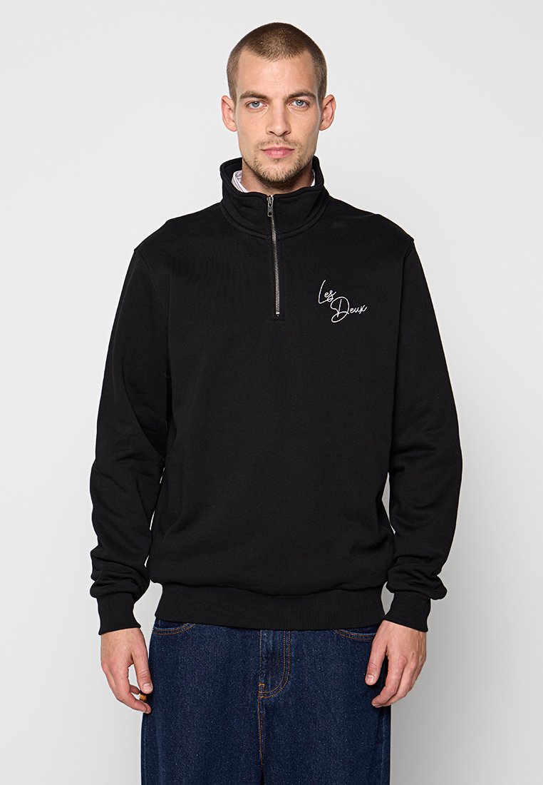 Mann in schwarzem Quarter-Zip-Pullover mit "Les Deux"-Schriftzug auf der Brust, kombiniert mit blauen Jeans, steht vor einem schlichten grauen Hintergrund.