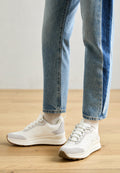 Weiße Sportschuhe mit grauen Akzenten und strukturiertem Obermaterial, kombiniert mit hellblauen, aufgeknöpften Jeans, vor einem Holzfußboden-Hintergrund.