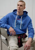 Blauer Baumwoll-Hoodie mit weißen Kordeln, mit "HUGO"-Logo und einem Stier-Grafik. Kombiniert mit cremefarbenen Hosen und einem schwarz-roten karierten Hemd.