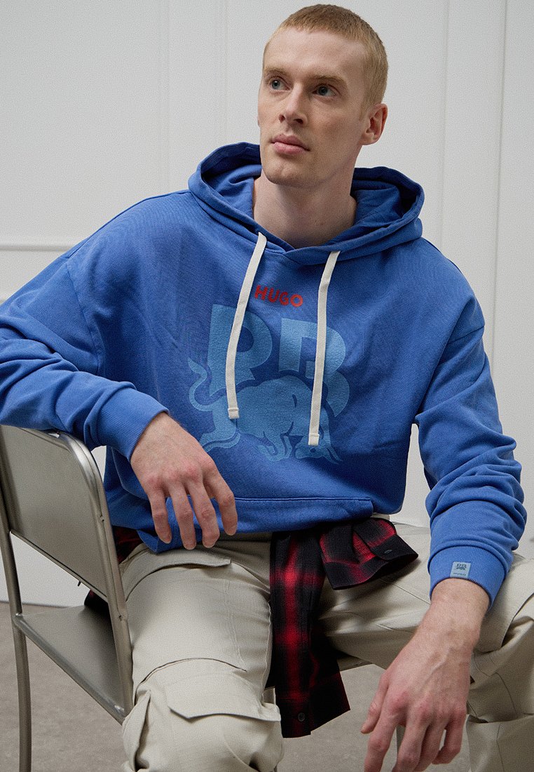 Blauer Baumwoll-Hoodie mit weißen Kordeln, mit "HUGO"-Logo und einem Stier-Grafik. Kombiniert mit cremefarbenen Hosen und einem schwarz-roten karierten Hemd.