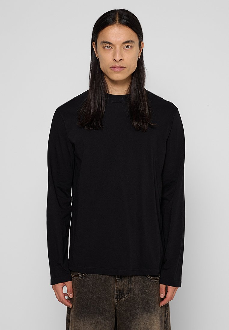 Junge Person mit langen dunklen Haaren, die ein schwarzes Langarmshirt und ausgebleichte dunkelblaue Jeans trägt, steht vor einem schlichten grauen Hintergrund.