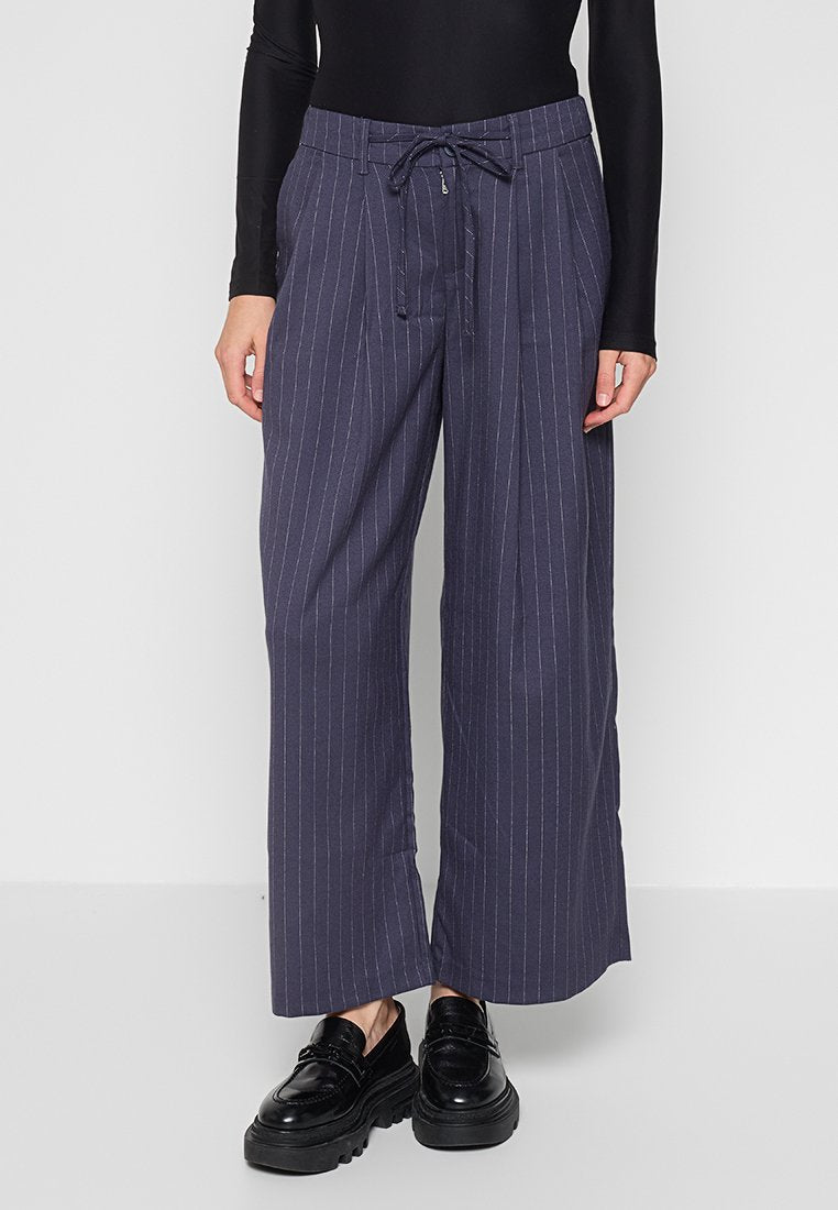 Modell trägt marineblaue, weit geschnittene Nadelstreifenhosen mit Kordelzug und schwarze klobige Loafers vor einem schlichten hellen Hintergrund.