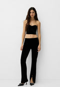 Schwarzes Samt-Crop-Top mit Herzausschnitt, kombiniert mit eng anliegenden schwarzen Hosen mit Schlitzen am Saum. Spitz zulaufende schwarze Absätze.