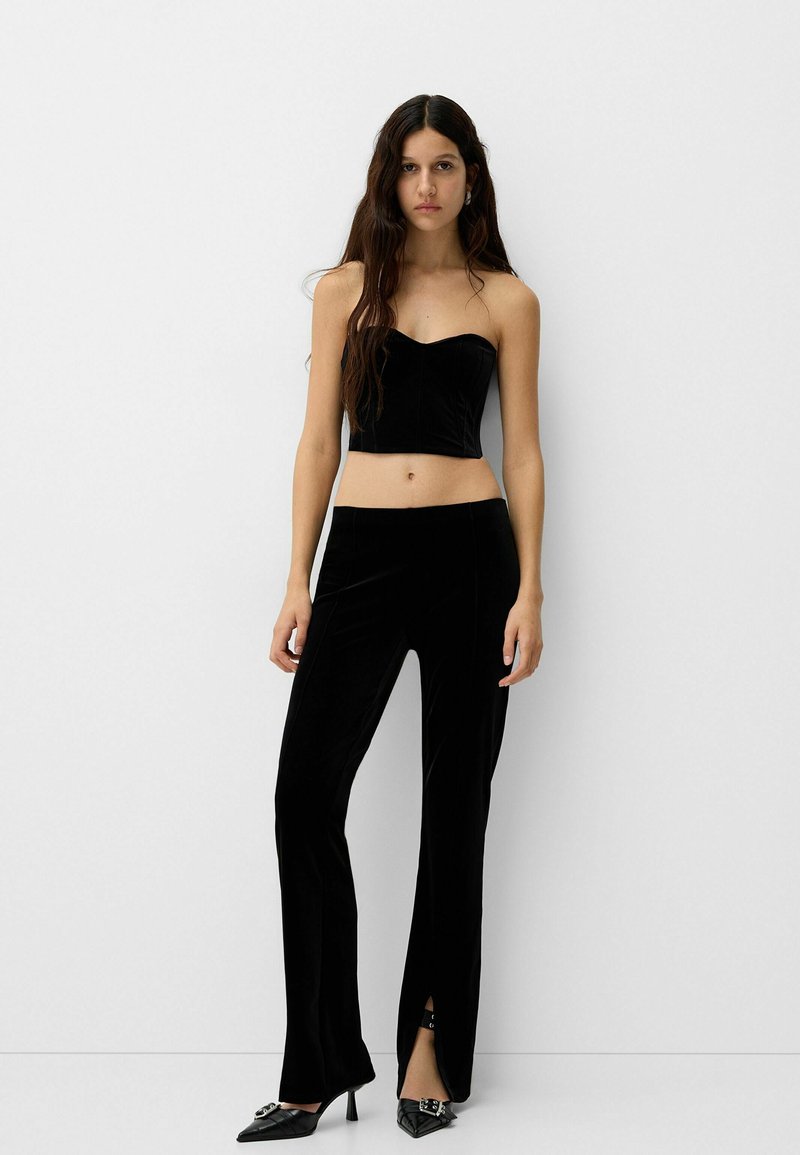 Schwarzes Samt-Crop-Top mit Herzausschnitt, kombiniert mit eng anliegenden schwarzen Hosen mit Schlitzen am Saum. Spitz zulaufende schwarze Absätze.