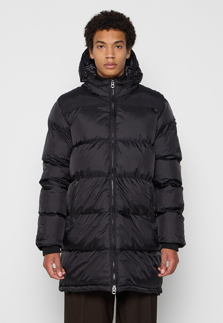 Junger Mann trägt eine schwarze wattierte Kapuzenjacke mit Frontreißverschluss und seitlichen Reißverschlusstaschen, steht vor einem schlichten hellen Hintergrund.