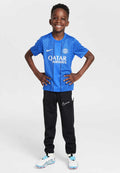 Blaue PSG-Trikot mit geometrischem Muster, schwarze Nike-Hosen und weiße Fußballschuhe mit blauen Akzenten. Kind lächelt und posiert selbstbewusst.