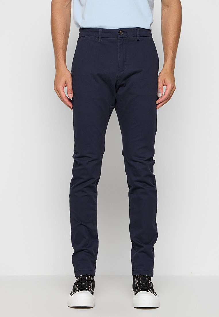 Marineblaue Chinos aus Baumwolle, mit schlanker Passform, vorderem Druckknopfverschluss und Seitentaschen. Getragen mit schwarz-weißen Turnschuhen.