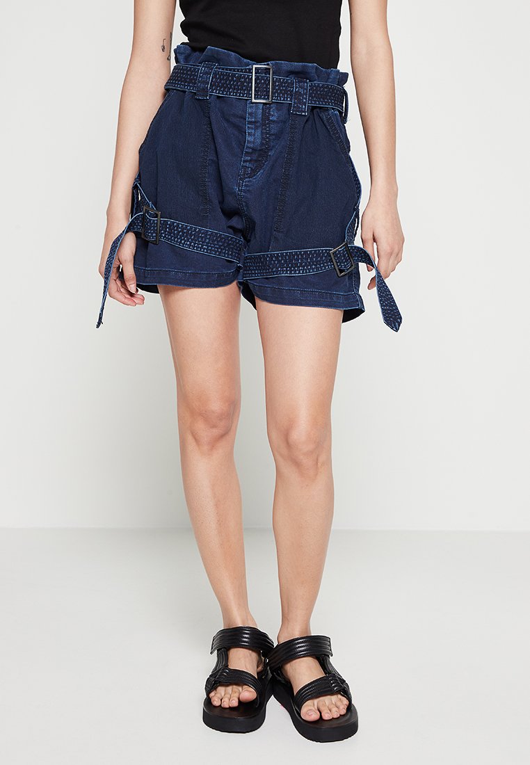 Blaue Denim-Shorts mit hoher Taille, verstellbarem Gürtel und Seitenschnürungen; verfügt über ein strukturiertes Muster und eine lockere Passform. Schwarze Plateausandalen.