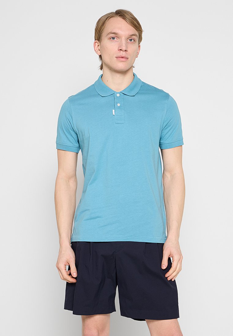 Hellblaues Poloshirt aus weichem Baumwollstoff, mit Kragen, zwei Knöpfen und kurzen Ärmeln. Kombiniert mit dunkelblauen Shorts.