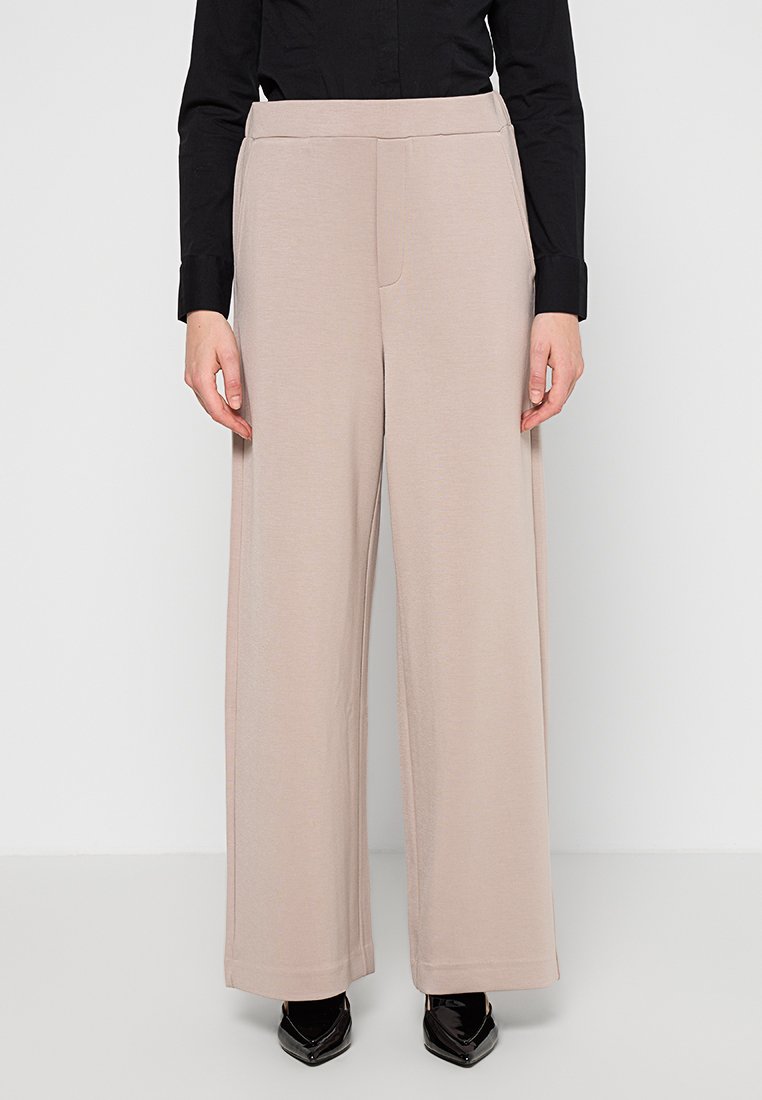 Beige weite Hosen aus weichem Stoff mit flachem Bund, hoher Taille und seitlichen Taschen, kombiniert mit schwarzen spitzen Schuhen.
