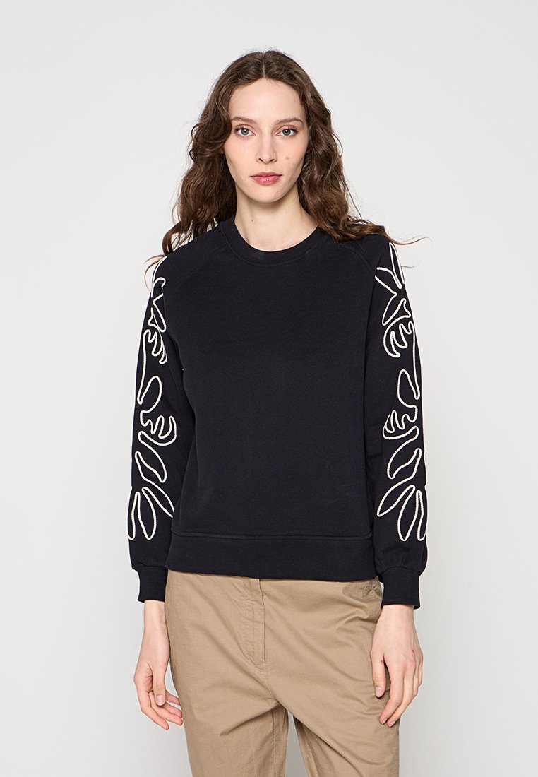Frau trägt schwarzen langärmligen Pullover mit weißen abstrakten Linienmustern auf den Ärmeln und beige Hosen, steht vor einem einfarbigen Hintergrund.