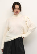 Cremfarbener Strickpullover mit hohem Kragen, lockerer Passform und strukturierten Ärmeldetails. Kombiniert mit einem schwarzen Lederrock.