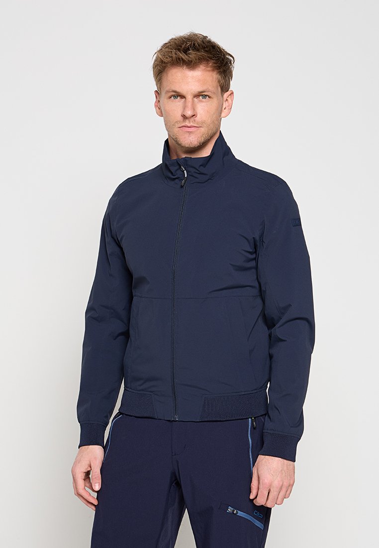 Mann trägt eine marineblaue Jacke mit Reißverschluss und hohem Kragen sowie passende marineblaue Hose mit Reißverschlusstasche.