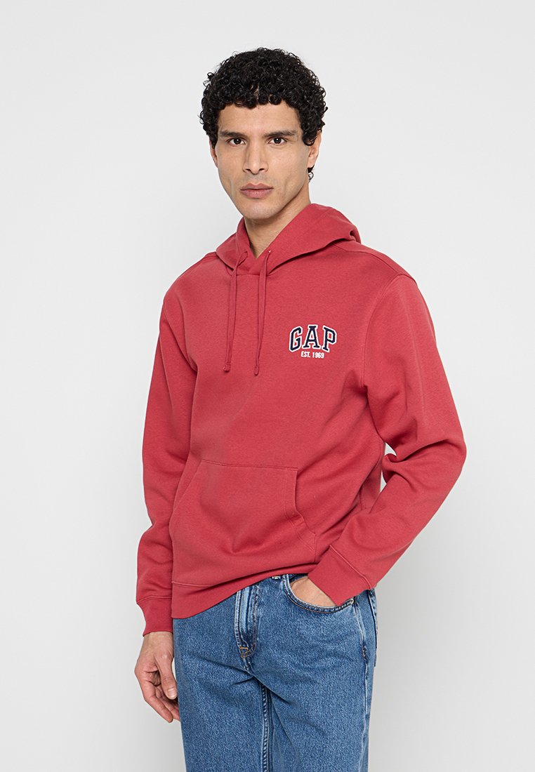 Mann mit lockigem Haar, der einen roten GAP-Kapuzenpullover und blaue Jeans trägt, steht vor einem schlichten hellen Hintergrund mit einer Hand in der Tasche.