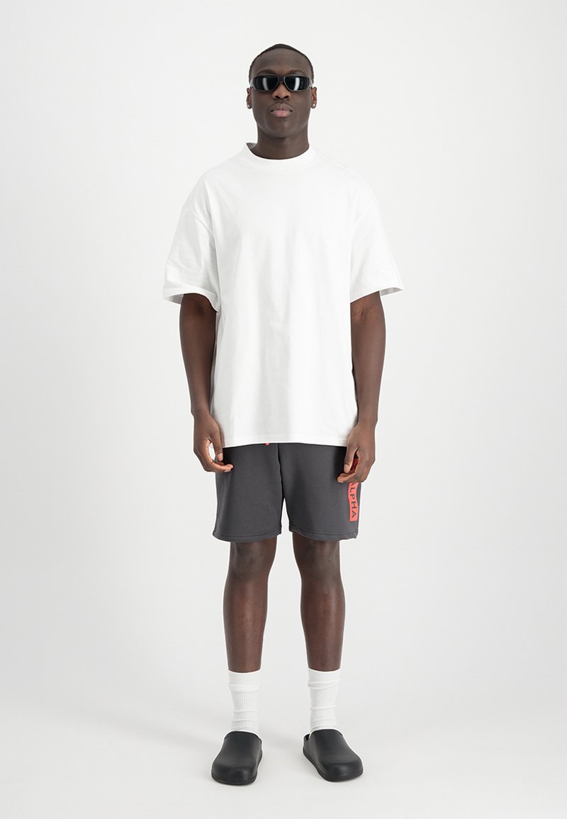 Weißes, übergroßes T-Shirt, graue Shorts mit rotem "ALPHA"-Text, weiße Crew-Socken, schwarze Slipper und schwarze Sonnenbrille.