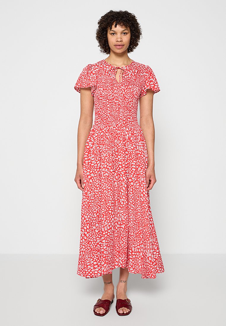 Frau steht und trägt ein rotes Midikleid mit Blumenmuster, Puffärmeln und burgunderroten Slip-On-Sandalen, vor einem einfarbigen hellen Hintergrund.