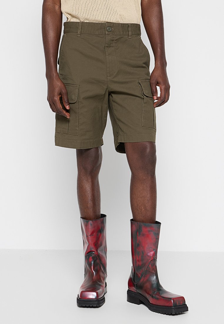 Khakifarbene Cargo-Shorts mit zwei großen Seitentaschen, mit geradem Schnitt und flachem Bund, kombiniert mit kniehohen rot-schwarzen Stiefeln.