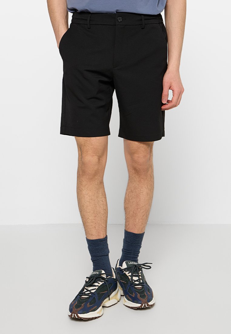 Schwarze Shorts aus elastischem Stoff, mit einem Knopfverschluss vorne und Seitentaschen. Getragen mit marineblauen Socken und gemusterten Turnschuhen.