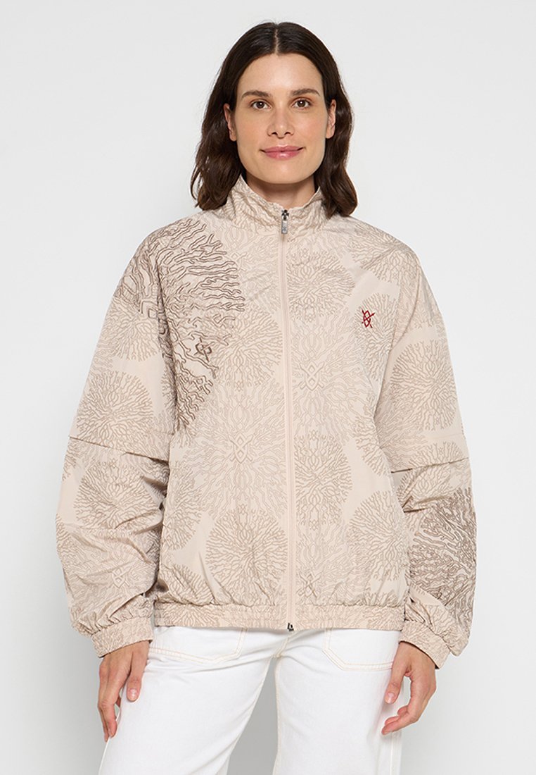 Beige Zip-Jacke mit strukturierten Mustern in dunklerem Beige, elastischen Bündchen und einem kleinen roten Logo auf der Brust.