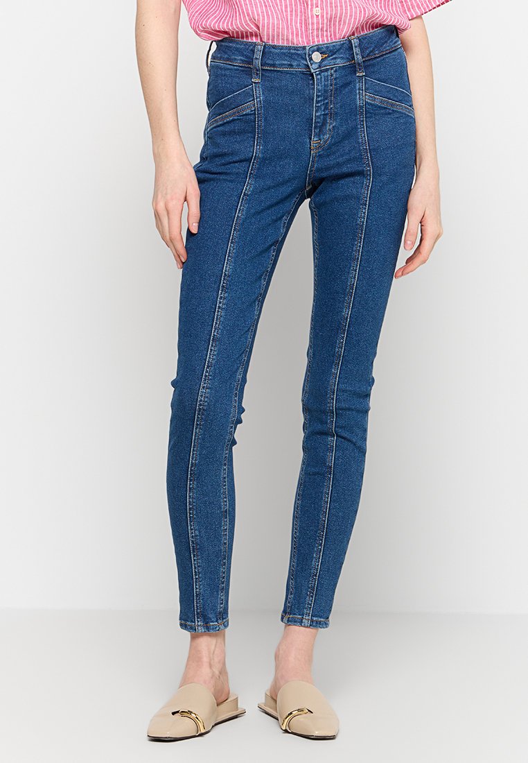 High-Waist blaue Skinny-Jeans aus Denim mit Vordertaschen und weißen Ziernähten, kombiniert mit beigen Slippern.