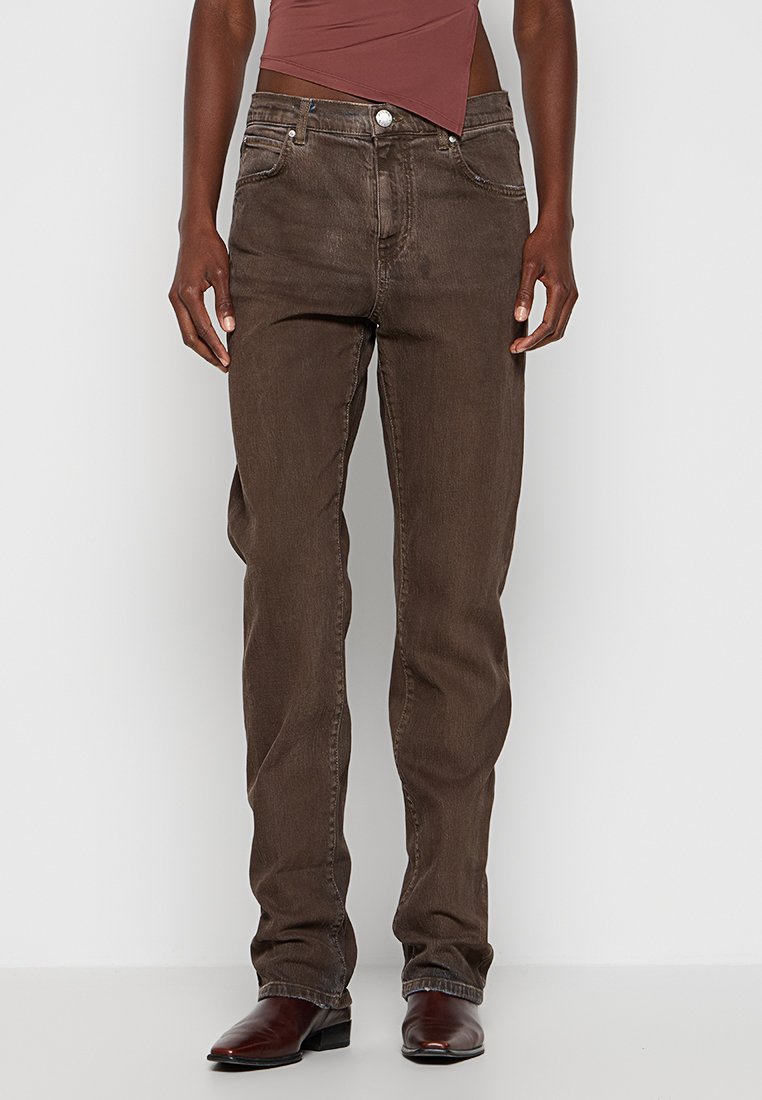 Person in gerad geschnittenen braunen Jeans, dunkelbraunen Lederstiefeln und einem asymmetrischen mauvefarbenen Oberteil vor einem schlichten hellen Hintergrund.