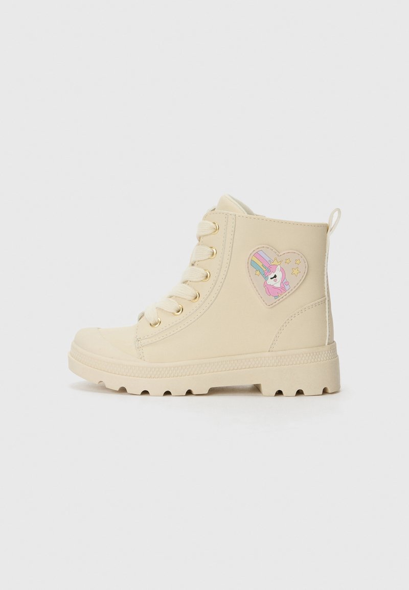 Beige Knöchelschuhe mit goldenen Ösen, strukturiertem Gummisohle und einem Einhornmotiv in Herzform auf der Außenseite.