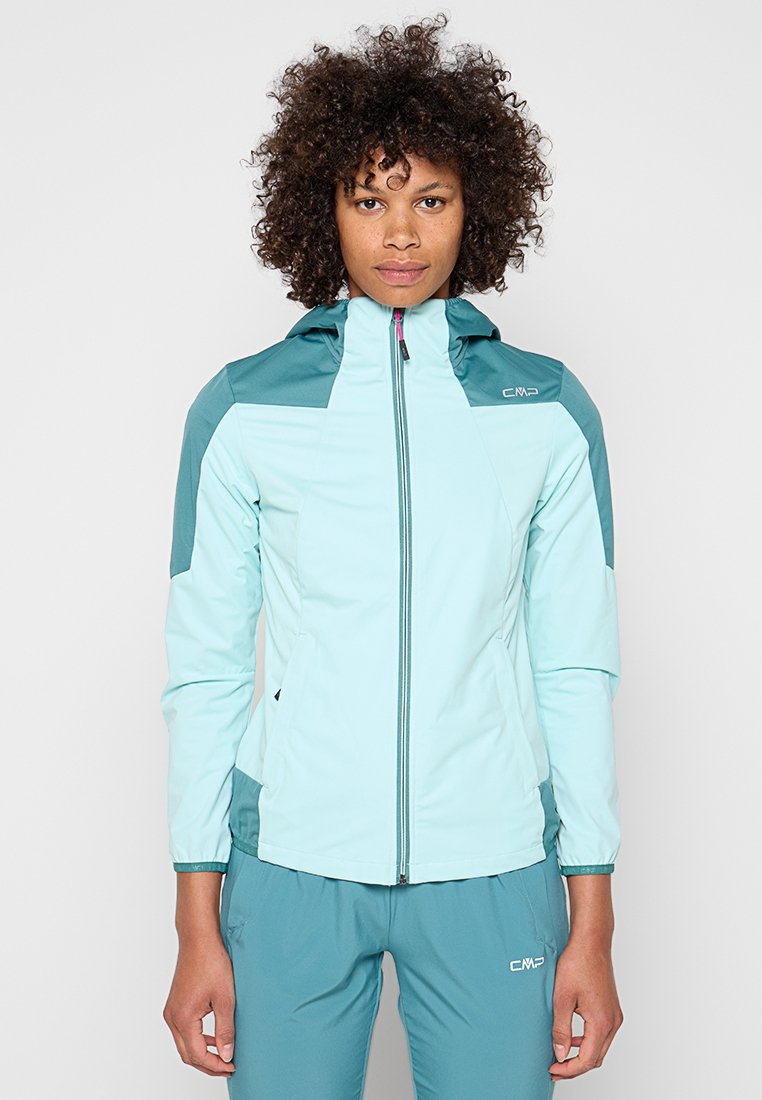 Frau mit lockigem Haar, die eine hellblaue und türkisfarbene Outdoor-Jacke mit Reißverschluss und passende türkisfarbene Hose trägt, steht vor einem einfarbigen Hintergrund.