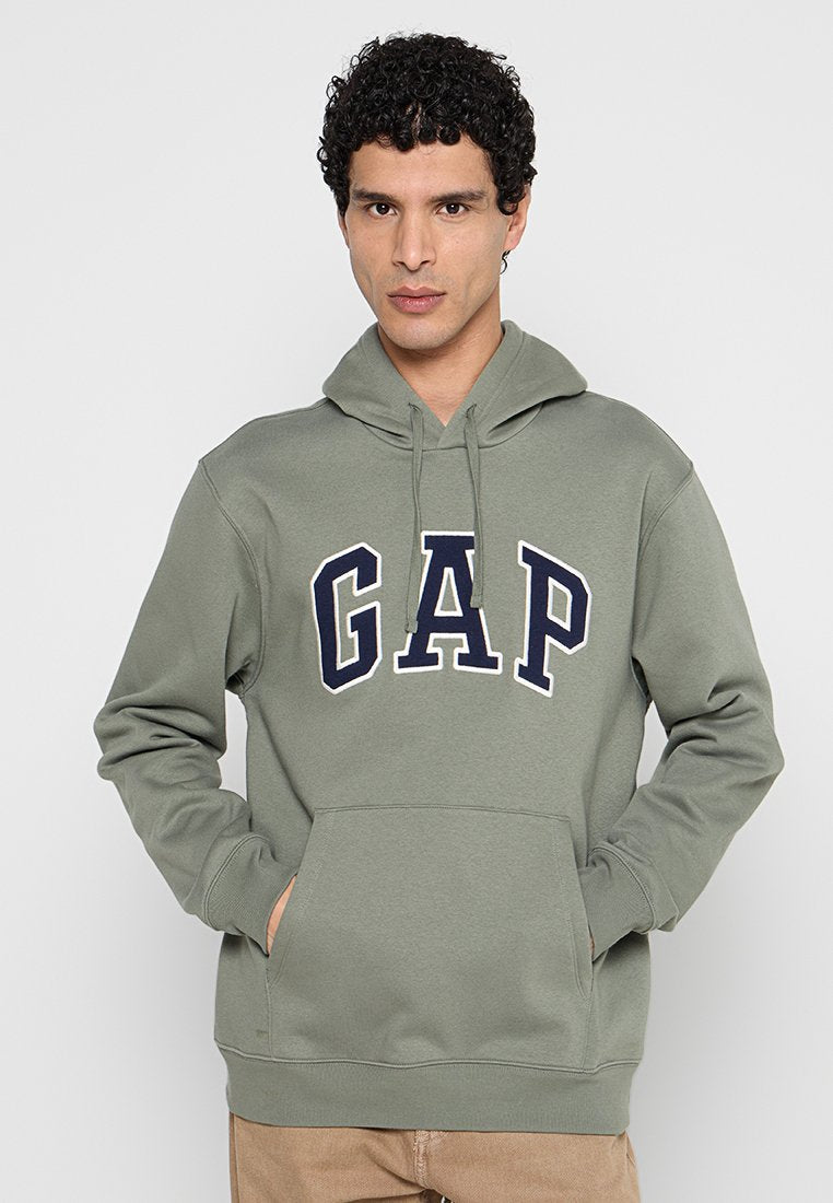 Mann trägt einen olivgrünen GAP-Hoodie mit den Händen in der vorderen Tasche, steht vor einem schlichten hellgrauen Hintergrund.