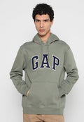 Mann trägt einen olivgrünen GAP-Hoodie mit den Händen in der vorderen Tasche, steht vor einem schlichten hellgrauen Hintergrund.