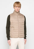 Beige Puffer-Weste mit vertikalen Nähten, hohem Kragen und frontalem Reißverschluss. Über einem dunkelblauen Langarmshirt getragen, das seitliche Taschen hat.