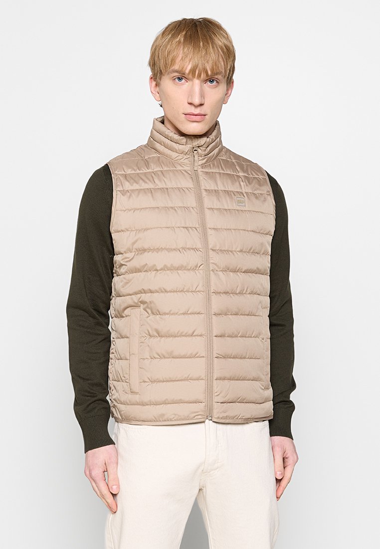 Beige Puffer-Weste mit vertikalen Nähten, hohem Kragen und frontalem Reißverschluss. Über einem dunkelblauen Langarmshirt getragen, das seitliche Taschen hat.
