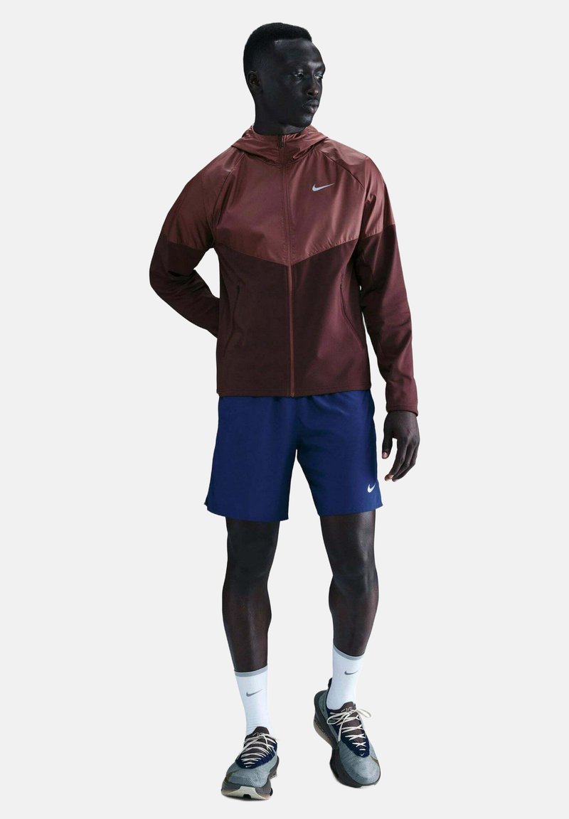 Mann trägt eine bordeauxrote Jacke mit Reißverschluss, marineblaue Shorts, weiße Nike-Socken und graue Laufschuhe, steht mit einer Hand hinter dem Rücken.