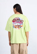 Person mit langen dunklen Haaren trägt ein übergroßes hellgrünes T-Shirt mit farbenfrohem, bonbon-inspiriertem Aufdruck und Text auf der Rückseite, beige Cargohose.