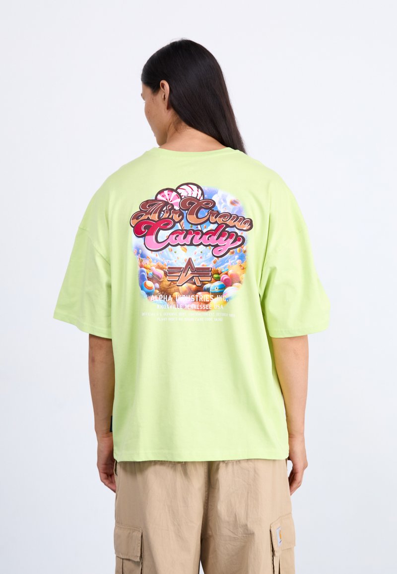 Person mit langen dunklen Haaren trägt ein übergroßes hellgrünes T-Shirt mit farbenfrohem, bonbon-inspiriertem Aufdruck und Text auf der Rückseite, beige Cargohose.