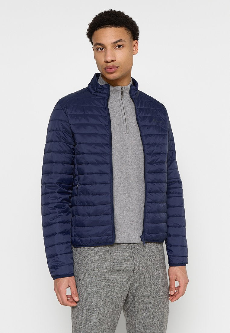 Marineblau gesteppte Pufferjacke mit Stehkragen und Reißverschluss. Der Träger trägt ein graues Zip-Hals-T-Shirt und graue Hosen.