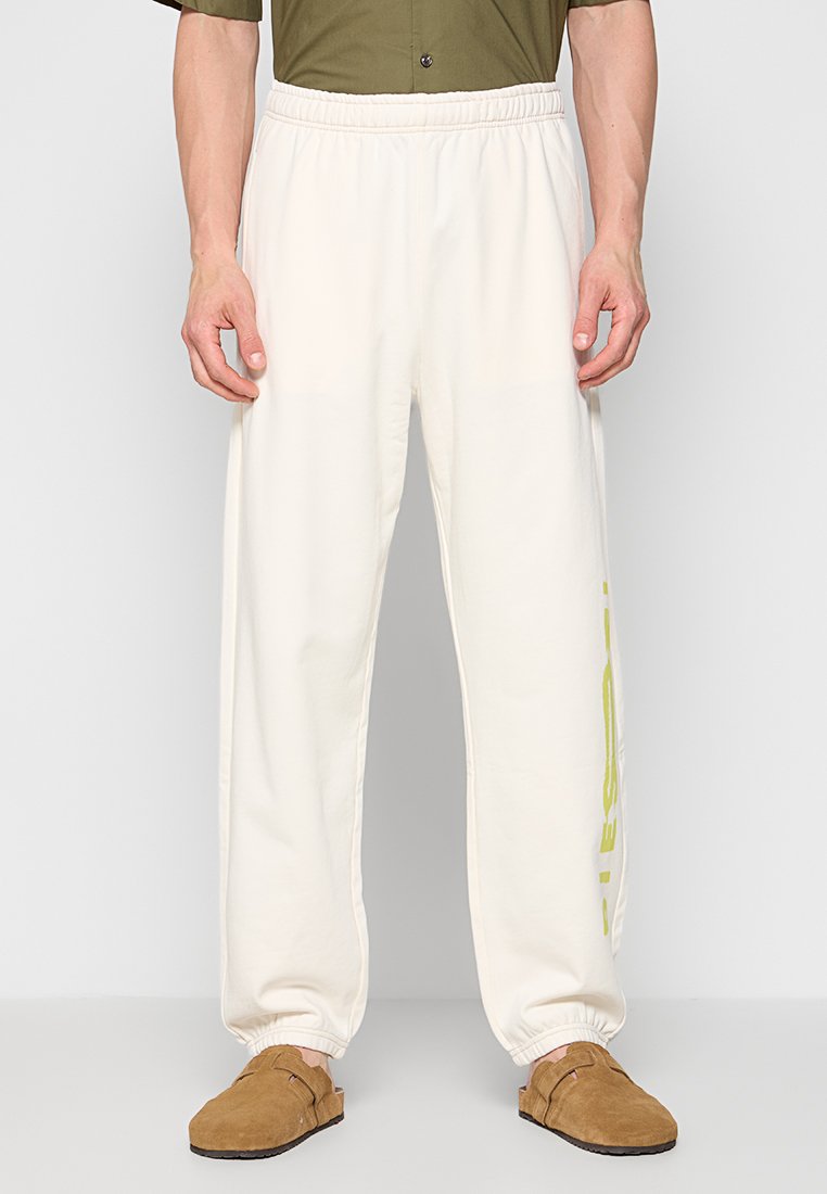 Mann in weißen Sweatpants mit elastischem Bund und gelbem vertikalem Text am linken Bein, stehend vor einem einfarbigen Hintergrund.