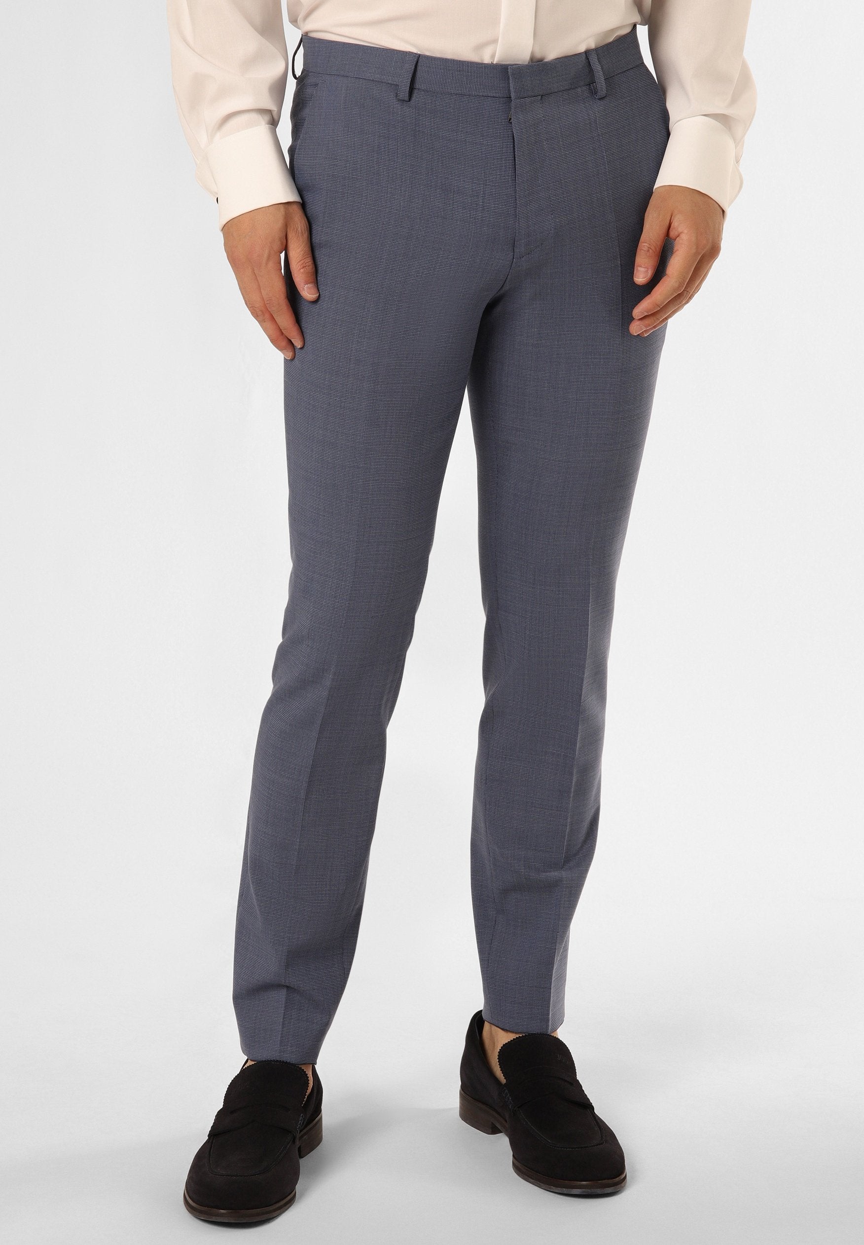 Mann in slim-fit grauen Anzughosen, schwarzen Loafers und einem hellbeigen langärmeligen Hemd, stehend vor einem weißen Hintergrund.