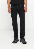 Mann trägt schwarze gerade geschnittene Jeans und schwarze Sportschuhe und steht vor einem schlichten weißen Hintergrund.