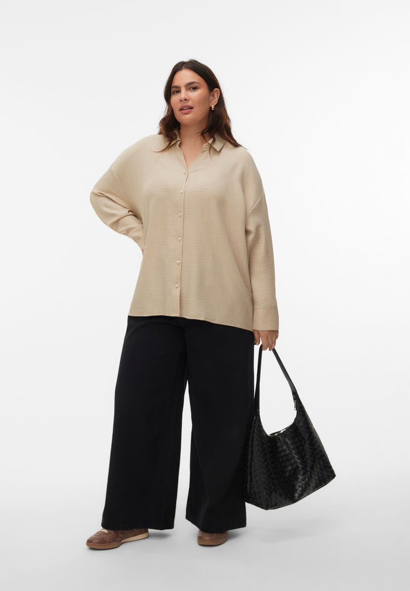 Beige Hemd zum Knöpfen mit lockerem Schnitt, kombiniert mit weit geschnittenen schwarzen Hosen. In einer Hand wird eine geflochtene schwarze Tasche gehalten, dazu braune Schuhe.