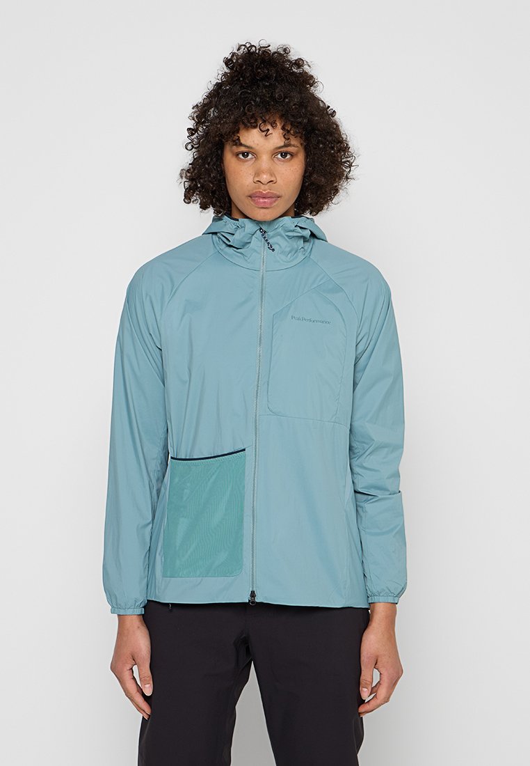 Junge Person mit lockigem Haar, die eine hellblaue Kapuzenjacke mit Frontreißverschluss und zwei Taschen trägt und vor einem schlichten Hintergrund steht.