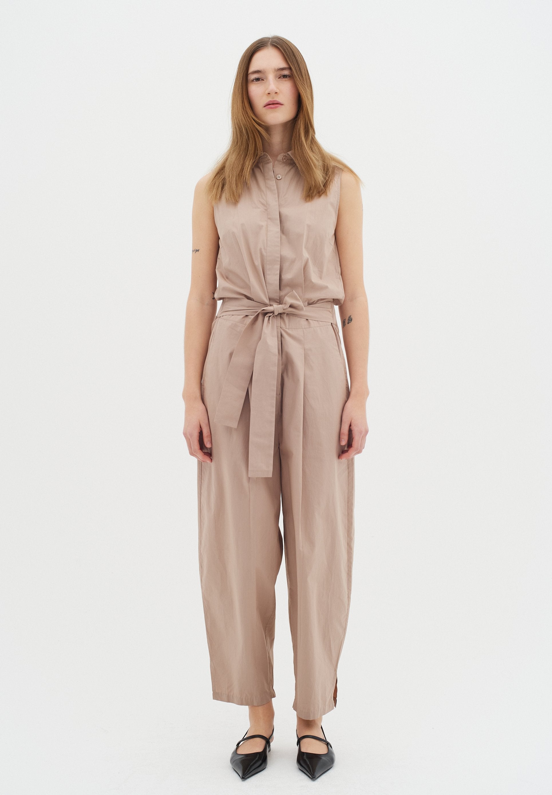 Beige ärmelloser Jumpsuit mit Kragen, tailliertem Schnitt, weitem Bein-Design und seitlichen Schlitzen. Kombiniert mit schwarzen spitzen Schuhen.