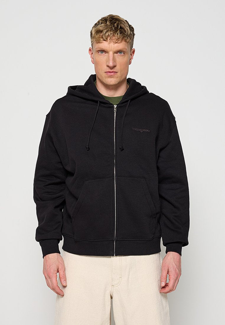 Blonder Mann trägt einen schwarzen Kapuzenpullover mit Reißverschluss und Fronttaschen sowie beige Hosen und blickt auf einem schlichten hellen Hintergrund nach vorne.