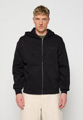 Blonder Mann trägt einen schwarzen Kapuzenpullover mit Reißverschluss und Fronttaschen sowie beige Hosen und blickt auf einem schlichten hellen Hintergrund nach vorne.