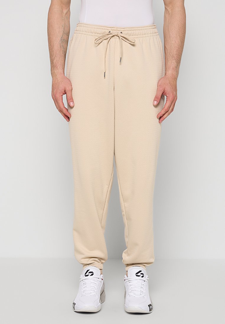 Beige Sweatpants aus strukturiertem Stoff mit elastischem Bund und Kordelzugdetail. Eng zulaufende Passform an den Knöcheln. Kombiniert mit weißen Sneakern.