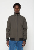 Mann trägt eine dunkel olivgrüne Zip-Jacke mit schwarzen Bündchen und Bund, kombiniert mit blauen Denim-Cargohosen, steht vor weißem Hintergrund.