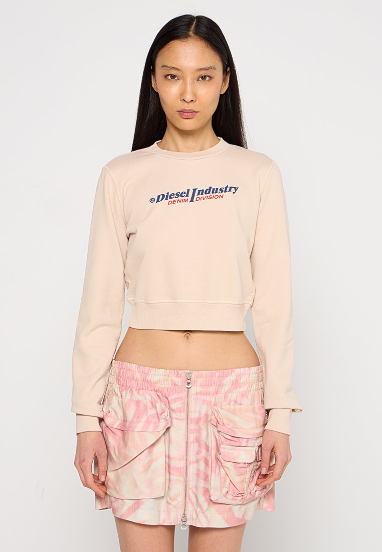Frau mit langen schwarzen Haaren, die einen beigen, verkürzten Sweatshirt trägt, auf dem "Diesel Industry" steht, und einen rosa gemusterten Rock mit großen Taschen.