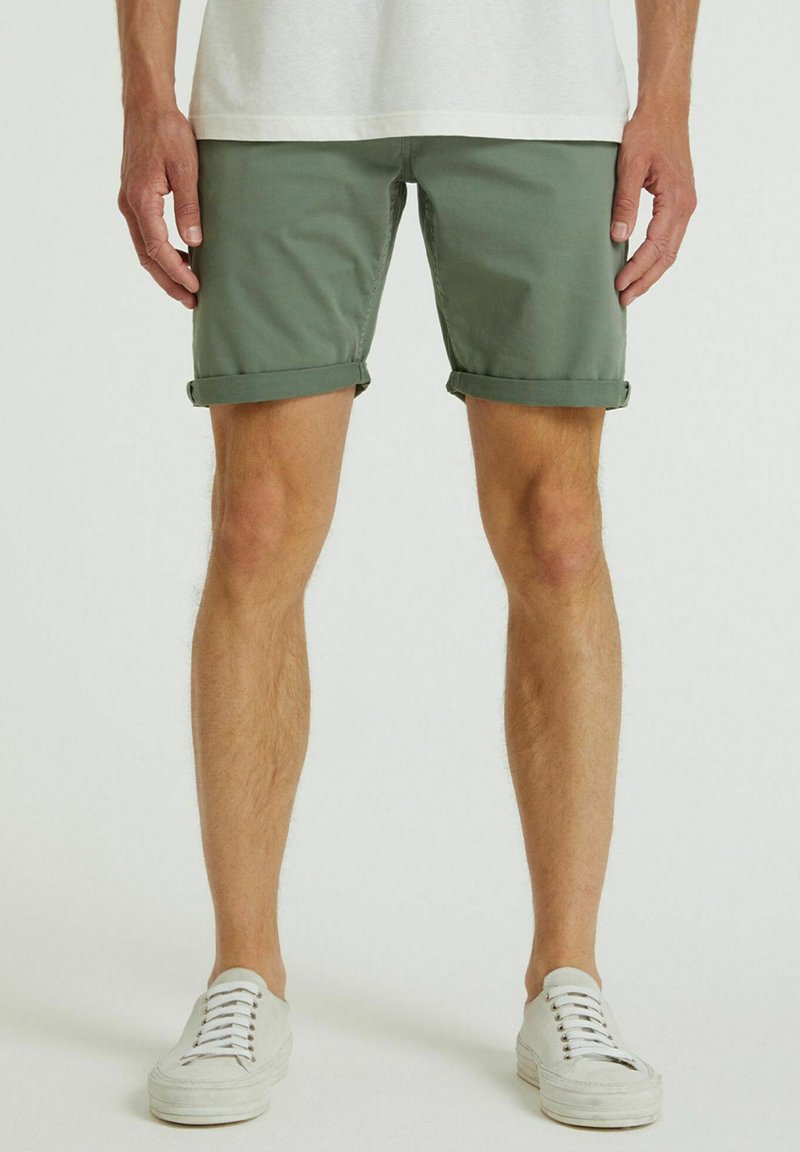 Olivgrüne Baumwollshorts mit umgeschlagenen Säumen, kombiniert mit weißen Schnürsneakers. Einfaches Design mit glatter Textur und taillierter Passform.